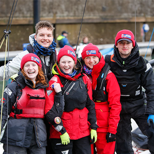 a group of young people wearing sailing standing on the shore and smiling at the camera