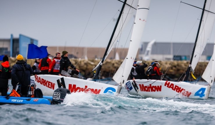 Two teams match racing Elliott 6M keelboats with the blue bulding of Weymouth and Portland National Sailing Academy in the background and an umpire on RIB waving a blue flag. 