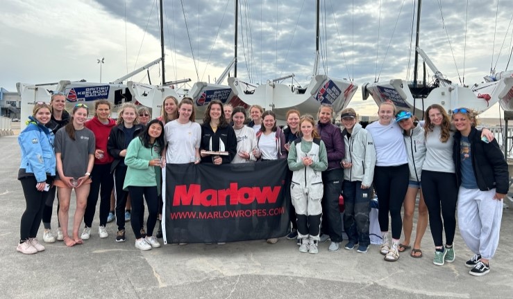 Group picture of 20+ smiling competitors on shore with keelboats behind at the RYA Marlow Ropes Women's Match Racing Championship 2024.